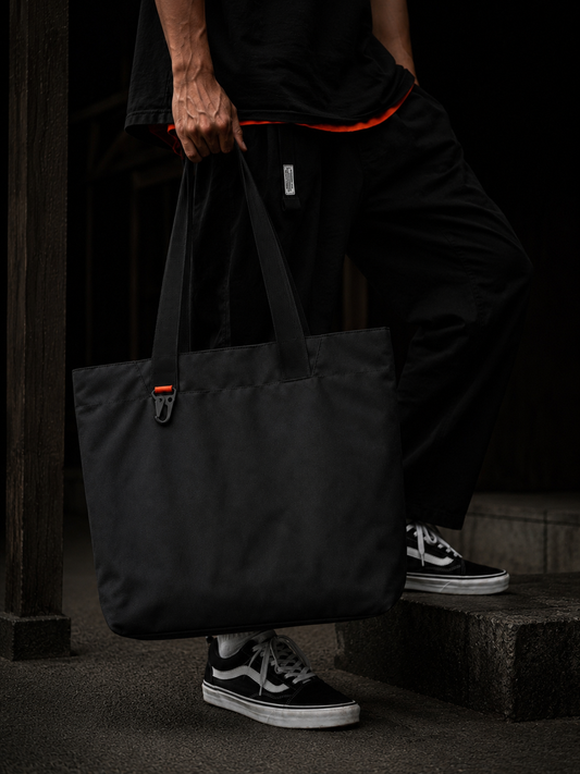 MAY.BE Utility Prime black tote, Drop 001, hanging against concrete wall